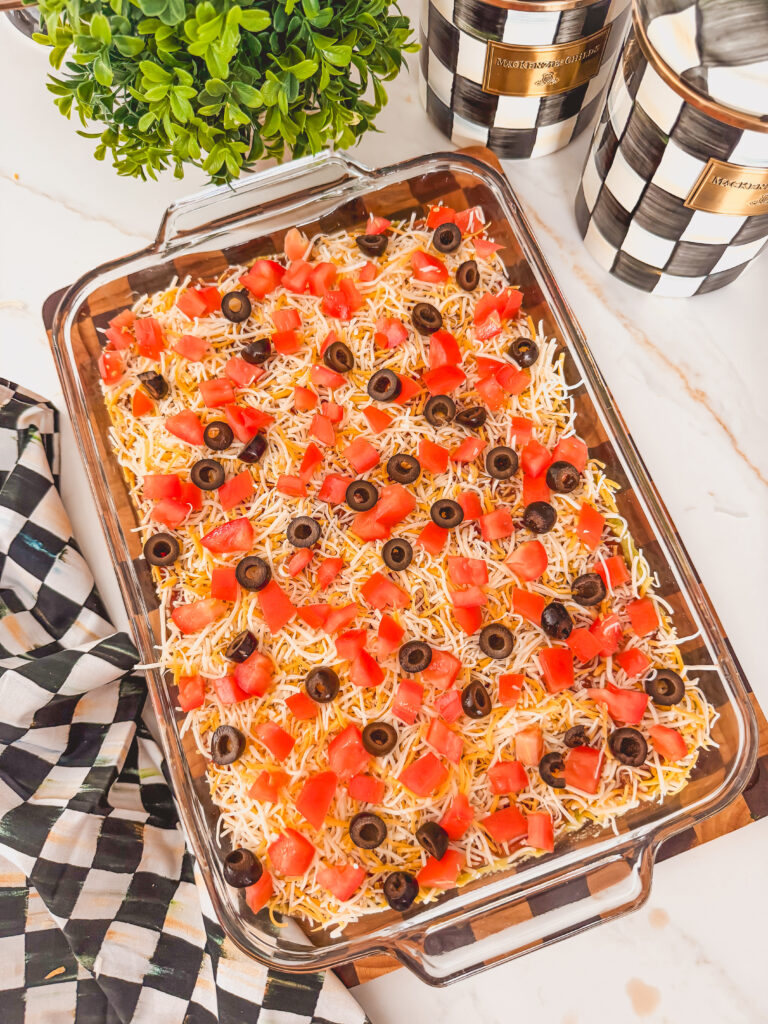 A glass baking dish with beans, guacamole, salsa, shredded cheese, tomatoes, and olives on top.