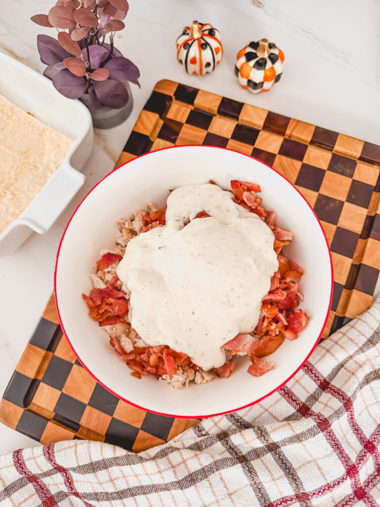 A white bowl with a red rim with shredded chicken, bacon, and ranch dressing on top.