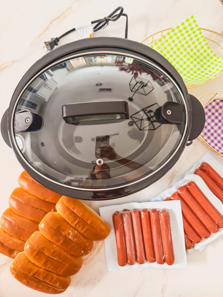 A crock pot with hot dogs and buns.