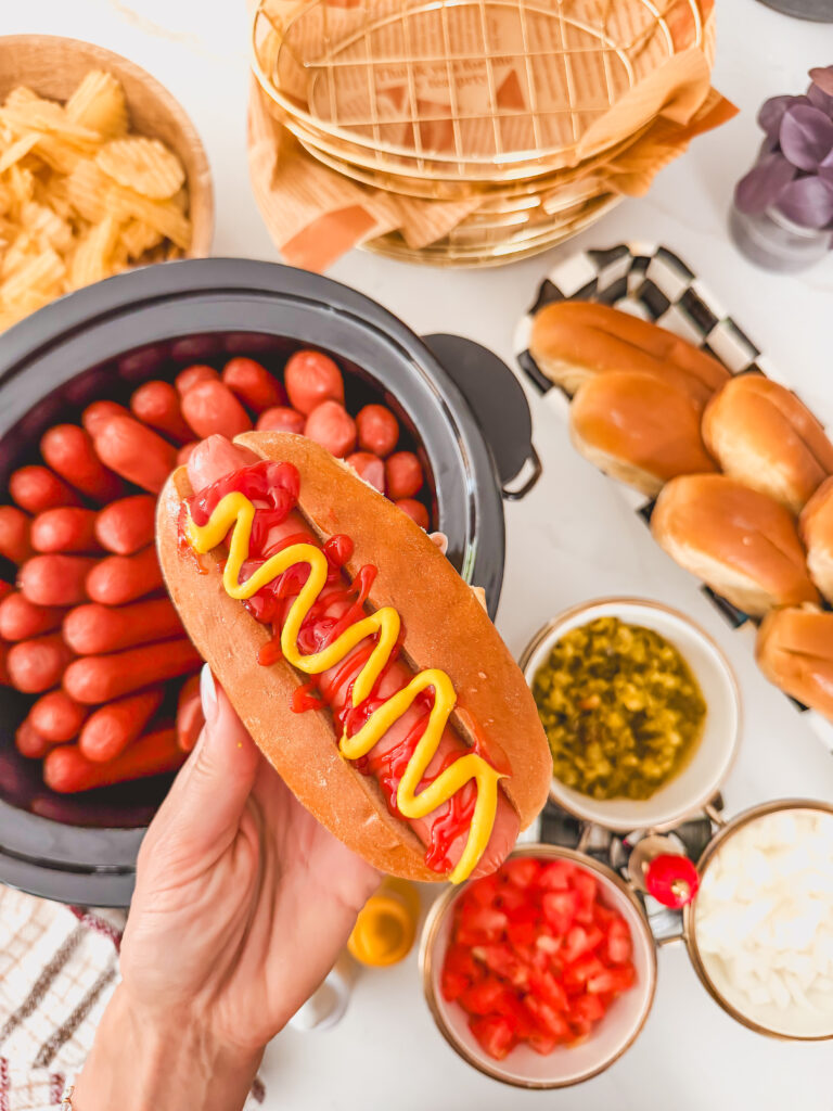Slow cooker hot dogs with a hand holding a hot dog and the slow cooker in the background.