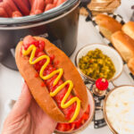 A hand holding a slow cooker hot dog with ketchup and mustard.