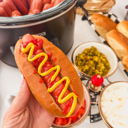 A hand holding a slow cooker hot dog with ketchup and mustard.