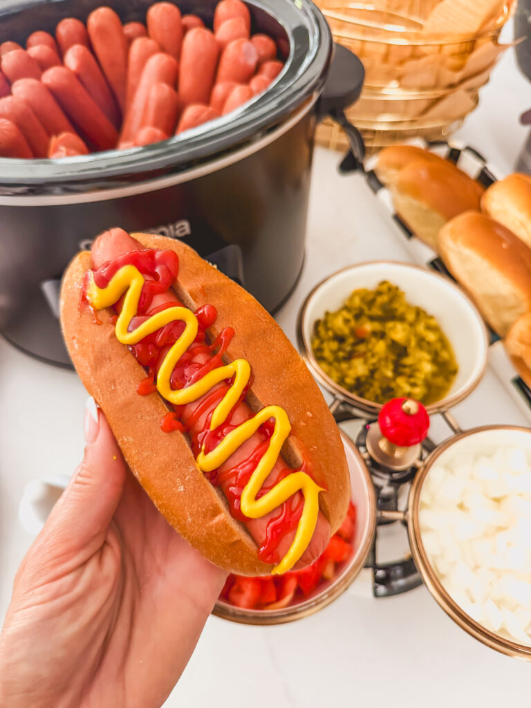 A hand holding a slow cooker hot dog with ketchup and mustard.
