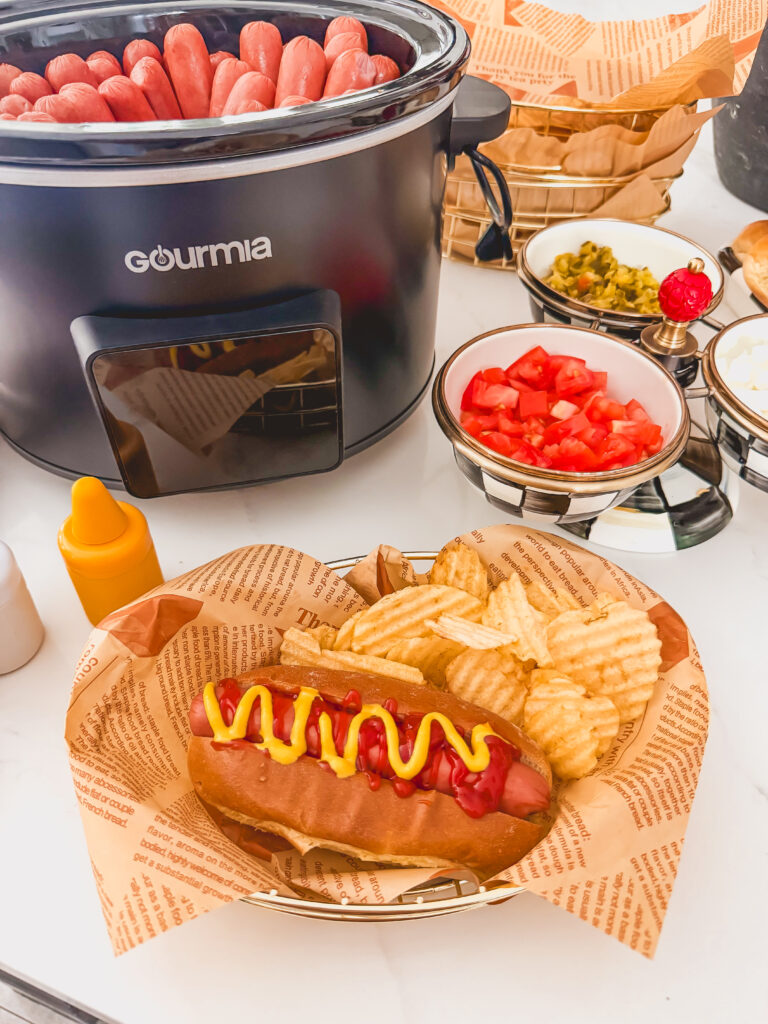 Slow cooker hot dog in a wire basket with chips and the slow cooker in the background.