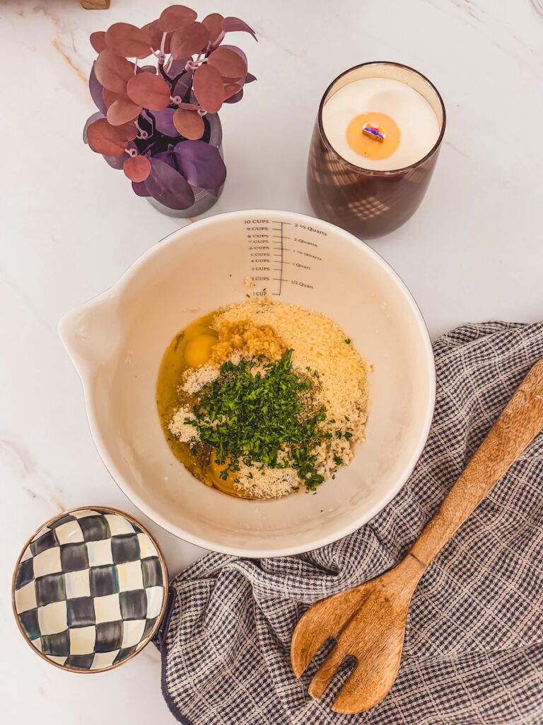 breadcrumbs, herbs and egg ingredients in white mixing bowl with spoon and dish towel next to it.