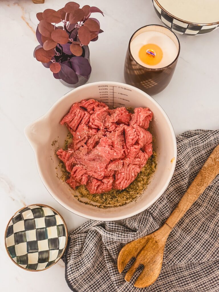raw ground meat in white bowl with other ingredients. wooden spoon and towel next to bowl.