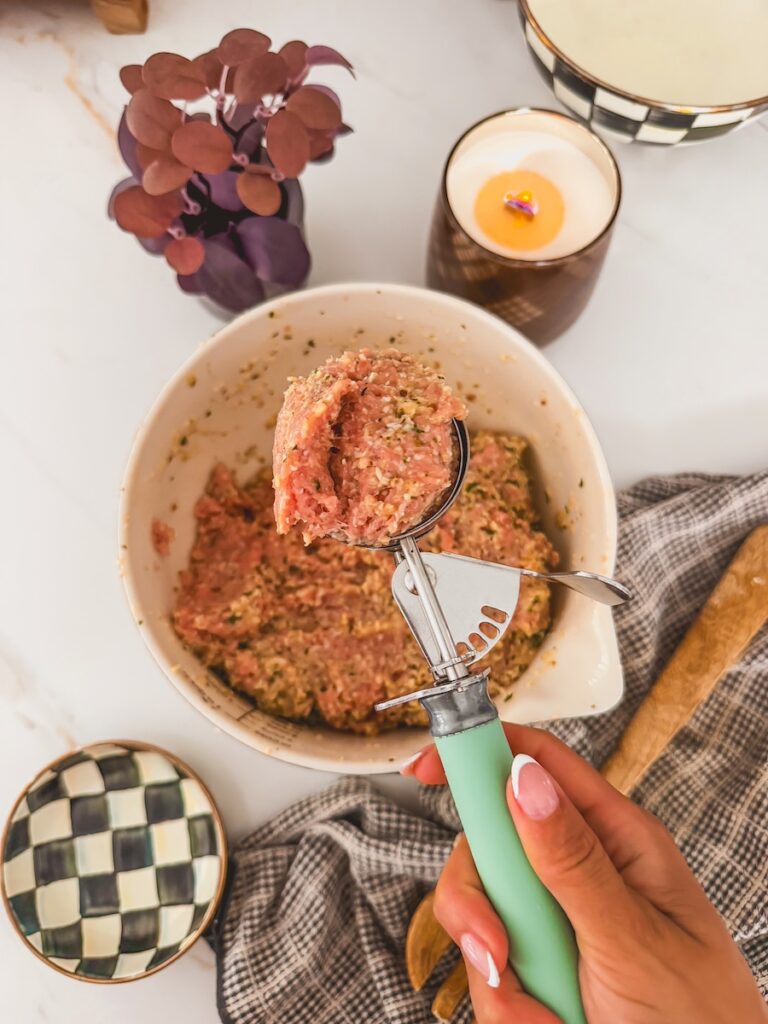 scoop of raw turkey meatball mixture with bowl of mixture beneath it. hand holding scoop.
