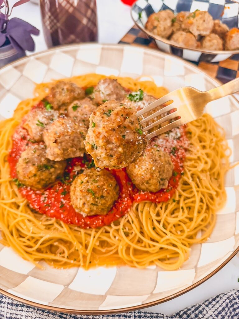 Gold fork with meatball on it with platter of meatballs, sauce, and pasta in background