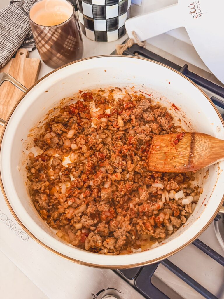 A white skillet with sausage, onions, tomato paste and a wooden spoon.