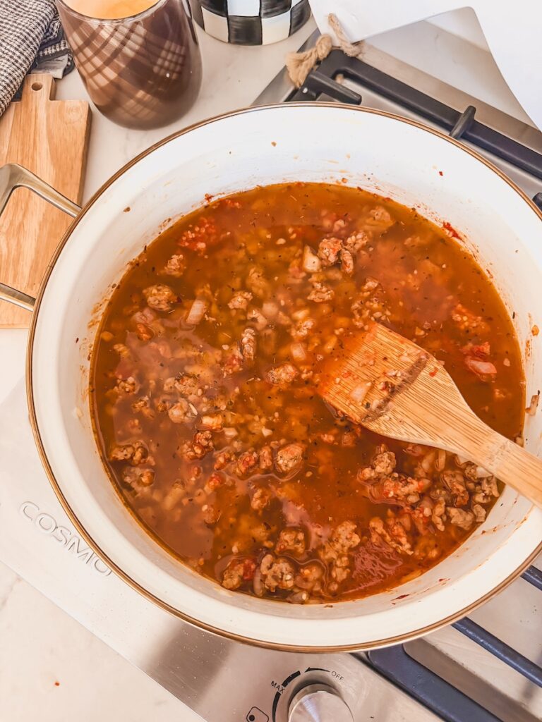 A skillet with a wooden spoon with chicken stock, sausage, spices, tomato paste, and spices.