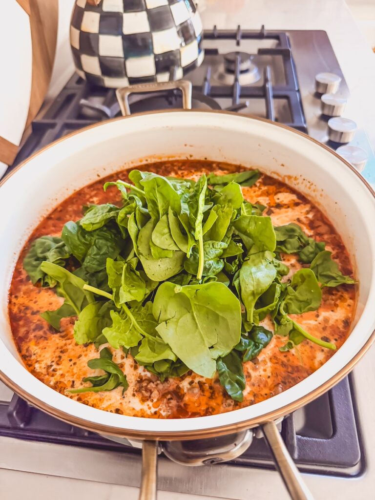 A white pot with ingredients for Italian sausage gnocchi soup with fresh spinach on top.