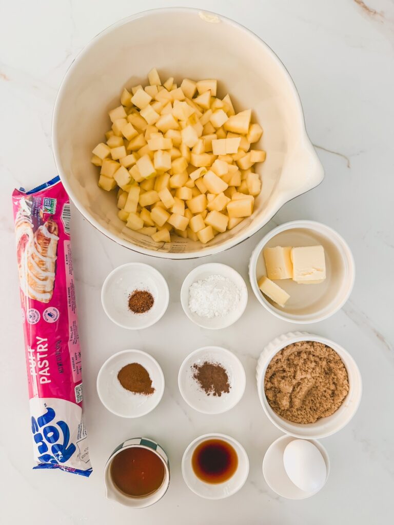 flatlay of ingredients including diced apples, puff pastry, cinnamon, maple syrup, nutmeg, all spice, brown sugar, butter, salt, vanilla
