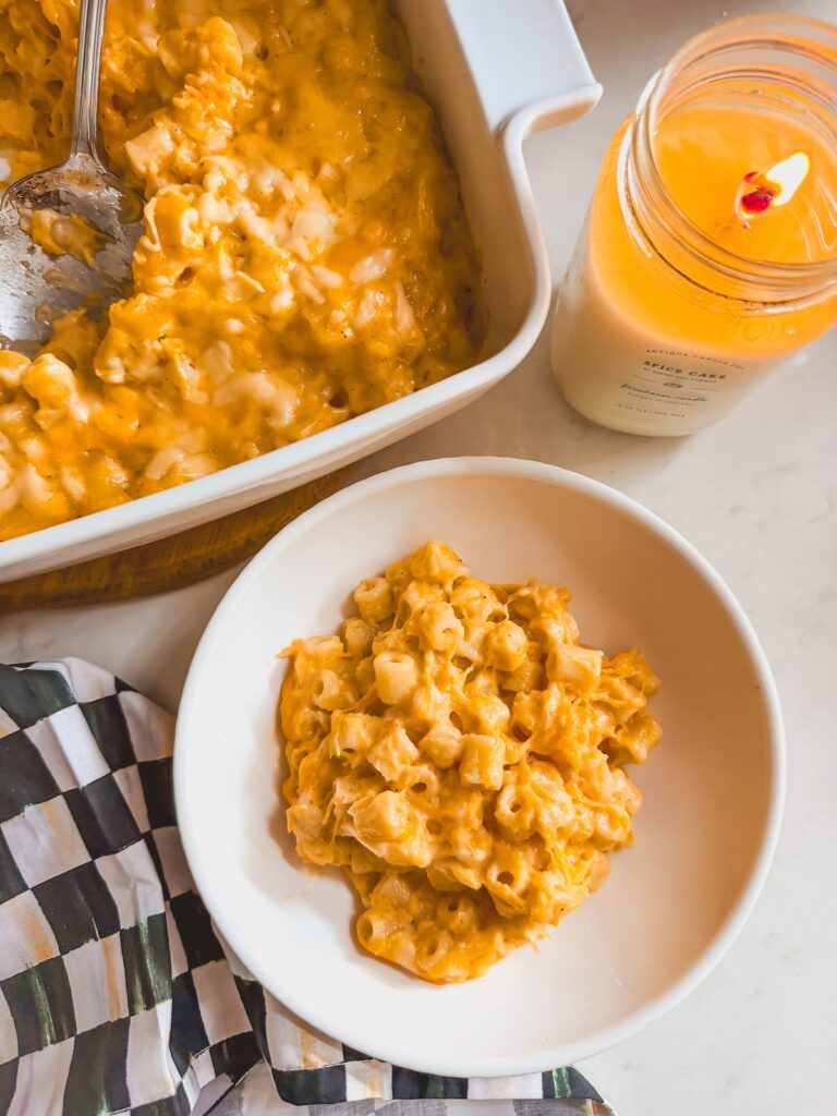 white bowl of mac and cheese next to baking dish of mac and cheese with a spoon. candle next to both.
