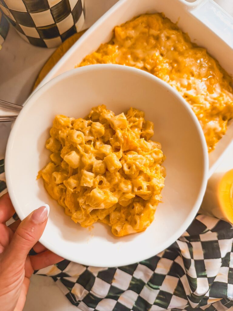 overhead shot of hand holding white bowl of mac and cheese with mac and cheese dish in background