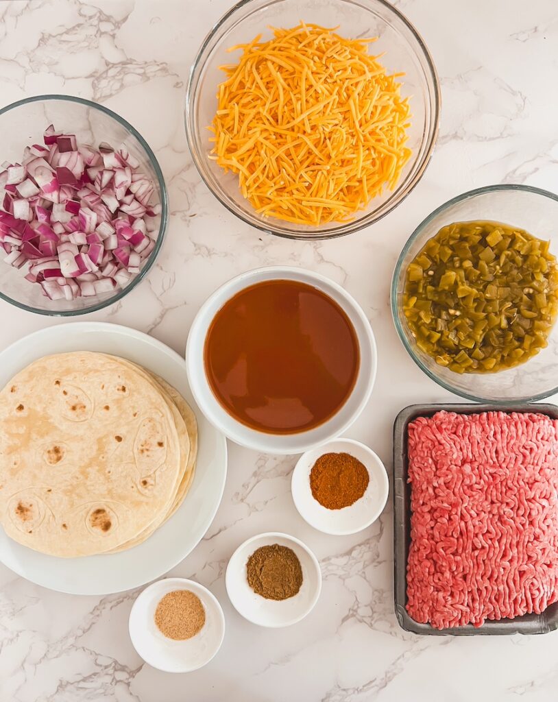 layered taco casserole flat lay of ingredients, beef, red onion, tortillas, green chilis, enchilada sauce, taco seasoning, cheddar cheese